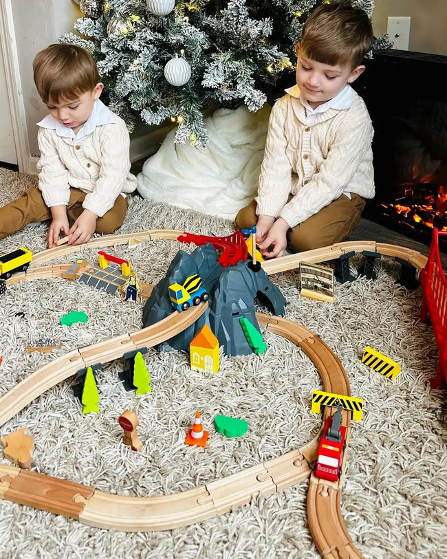 Two children playing with a toy train set in front of a Christmas tree.