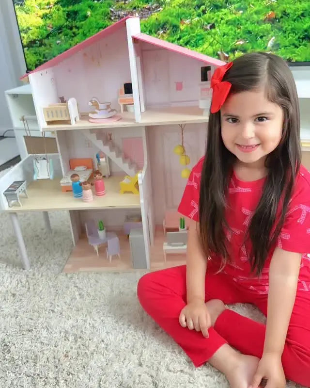 Young girl in red pajamas sitting next to a dollhouse with a forest background