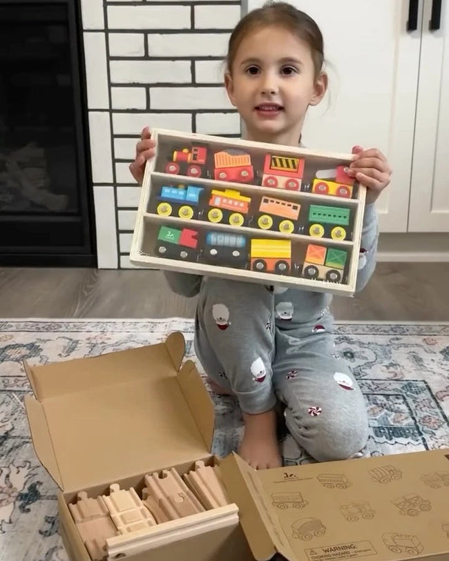 Child holding a toy train set box with wooden tracks on a living room floor.